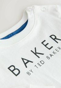 Biela bavlnená tričko s modrým vnútorným golierom, s veľkým čiernym textom "BAKER" a menším textom "by TED BAKER." Viditeľné sú dve cvoky.