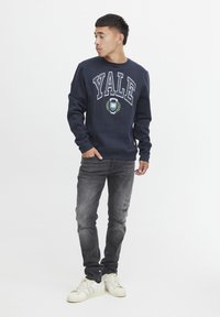 Felpa navy con la scritta "YALE", caratterizzata da un logo e un design di alloro. Abbinata a jeans grigi sbiaditi e sneakers bianche. Stile casual.