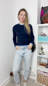 Jūras spēku adīta džemperis ar gaiši zilu apkakli, gaiši zili džinsi un baltas kedas. Melna soma; stāvot pie baltām sienām ar plauktiem.