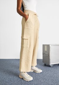 Osoba nosząca beżowe, teksturowane spodnie cargo z bocznymi kieszeniami i sneakersami, stojąca obok srebrnej metalowej walizki na kółkach.