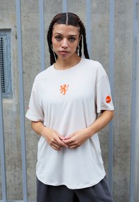 Bež oversized majica s oranžnim grafičnim motivom levjega na prsih in Nike logotipom na rokavu. Mehka tkanina z okroglim izrezom in kratkimi rokavi.
