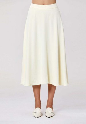 Jupe midi taille haute de couleur crème, associée à des chaussures blanches à bouts pointus sans lacets, portée par une personne debout devant un fond uni.