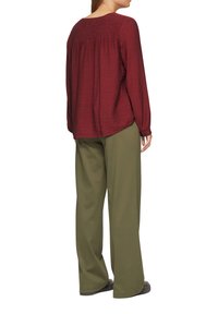 Blouse longue maroon texturée avec dos froncé et coupe ample, associée à un pantalon large olive. Chaussures noires visibles.