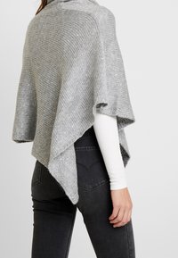 Femme portant un poncho gris en tricot avec un ourlet diagonal sur un haut à manches longues blanc et un jean noir, vue de dos et de côté.