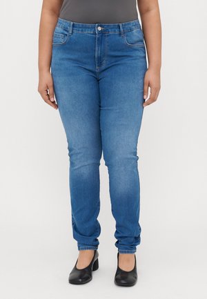Frontbillede af en person iført mellemblå slim-fit jeans, sorte sko med lav hæl og en mørkegrå indstukket top mod en ensfarvet baggrund.