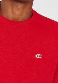 Sweat-shirt rouge avec encolure côtelée et tissu texturé. Présente un petit logo blanc sur le devant, mettant en valeur le design décontracté.