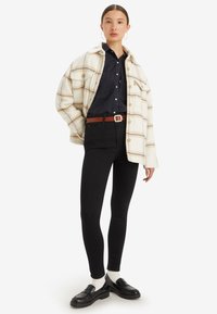 Mladá žena oblečená v béžovej károvej bunde, čiernom tričku, čiernych obtiahnutých džínsach, bielych ponožkách, čiernych mokasínach a kruhových náušniciach, stojaca proti bielej stene.