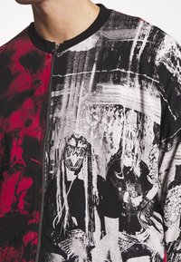 Zip-up jacket featuring a black and red color scheme, abstract designs, and graphic prints of figures with masks. Textured fabric.