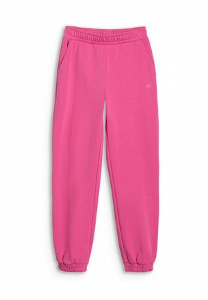 Fille debout les mains dans les poches, portant un haut blanc à manches longues, un pantalon de jogging rose et des baskets noires sur fond uni.