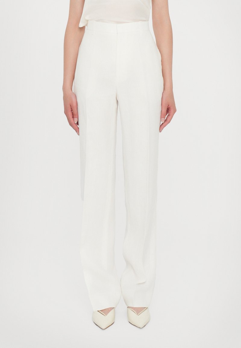 White, high-waisted trousers with a wide leg silhouette, featuring a smooth texture and tailored fit, paired with pointed-toe heels.