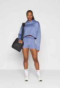 Hoodie cropped azzurro chiaro con cordini e logo "NIKE PRO", abbinato a pantaloni corti coordinati. Una borsa nera e scarpe bianche Nike completano il look.