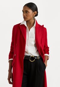 Palton roșu din lână cu un design cu guler dublu, cămașă albă cu guler și dungi verticale, curea neagră cu o cataramă circulară aurie și pantaloni negri.