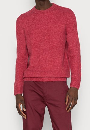 Mann trägt einen langärmligen roten Strickpullover und dunkelrote Hose, steht vor einem einfarbigen hellen Hintergrund.