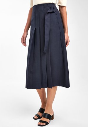 Femme portant une jupe midi plissée bleu marine avec une ceinture sur le côté, associée à des sandales à talons noires et un haut beige clair.