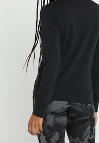 Sweat-shirt noir à manches longues, texture lisse et coupe décontractée. Associé à des leggings gris à motifs avec des formes abstraites.