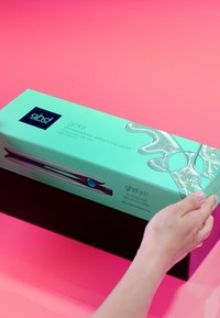 Main tenant une boîte vert menthe d'un lisseur professionnel avancé ghd Gold Silver sur un fond rose.