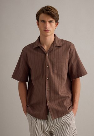 Giovane uomo con capelli castani corti indossa una camicia marrone a maniche corte con bottoni e pantaloni bianchi con sottili strisce verticali nere.