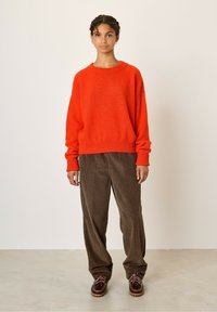 Rdeča volnena pulover s sproščenim krojem, rebrastimi manšetami in robom. V kombinaciji z rjavimi kordelnimi hlačami in temno rjavimi vezalkami. Preprost ozadje.