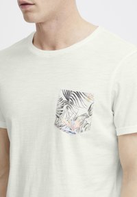 Camiseta blanca de tela suave con un bolsillo en el pecho que presenta un estampado de hojas tropicales de colores. Cuello redondo y mangas cortas con puños doblados.