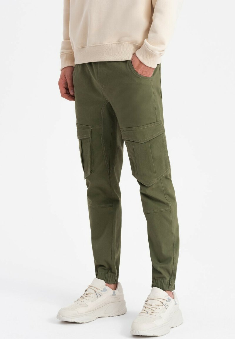 Olivgröna cargobyxor med flera sidofickor, avsmalnande anklar och en bomullsblandning, bärs med en ljusbeige sweatshirt och sneakers.
