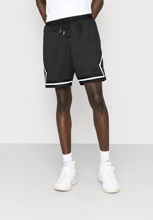 Homme portant un short de sport en mesh noir avec bordure blanche, des baskets blanches et des chaussettes blanches, debout devant un fond clair uni.