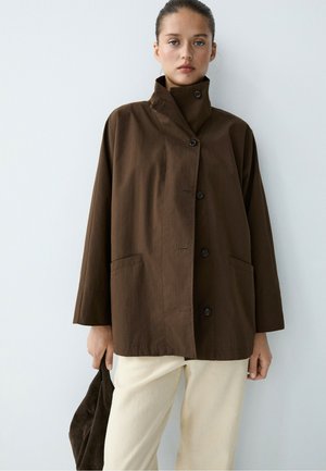 Femme portant une veste boutonnée marron foncé à col montant et un pantalon beige clair, tenant un sac marron foncé assorti sur fond blanc.