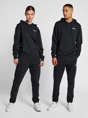 Zwei Models tragen schwarze Kapuzenpullover und passende Jogginghosen mit weißen Sneakern und stehen vor einem einfarbigen hellen Hintergrund.