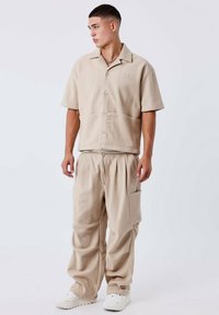 Camicia beige a maniche corte con bottoni, due tasche frontali e pantaloni larghi abbinati con vita elastica e tasca laterale.