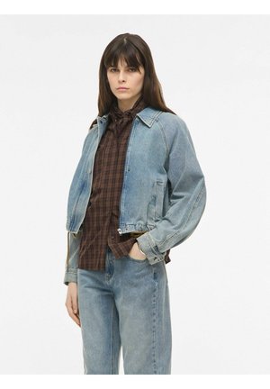 Giovane donna con lunghi capelli castani indossa una giacca di jeans azzurro chiaro e jeans sopra una camicia marrone a quadri, posando con una mano in tasca.