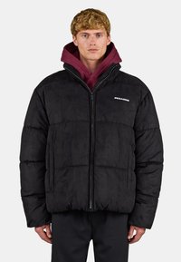 Jeune homme aux cheveux blonds courts et bouclés, portant une doudoune noire sur un sweat à capuche bordeaux, debout devant un fond clair uni.