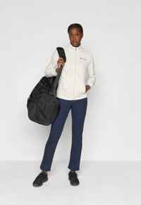 Chaqueta de forro polar blanca con cremallera y logo, pantalones azul marino y bolsa de deporte negra. Zapatillas deportivas negras con suelas texturizadas. Fondo blanco liso.
