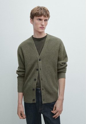 Jeune homme portant un cardigan côtelé olive sur une chemise vert foncé et un jean bleu marine, se tenant devant un fond clair et uni.