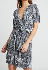 Vestido de estampado de serpiente en blanco y negro, con mangas cortas, un escote en V profundo y un detalle de cintura anudada. Presenta una falda fluida.