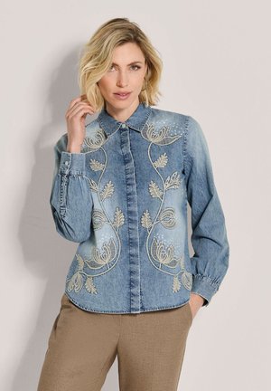 Vrouw met blond haar die een lichtblauw spijkerblouse met geborduurde bloemenpatronen en beige broek draagt, staand tegen een effen achtergrond.