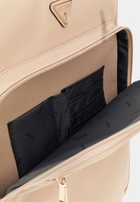 Interno di uno zaino Beige Guess che mostra una fodera nera con un motivo di logo ripetuto, diversi slot per schede e una zip dorata.