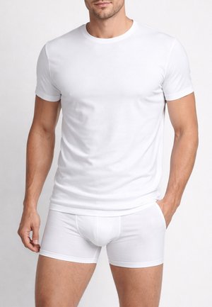 Uomo che indossa una semplice maglietta bianca a maniche corte e boxer bianchi abbinati, in piedi con una mano in tasca.