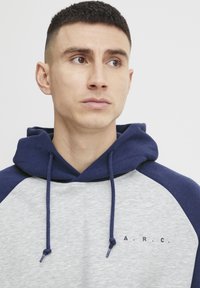 Hoody met een grijze body en marineblauwe raglanmouwen. Voorzien van trekkoorden en "A.R.C." gedrukt op de borst. Zachte, casual stof.