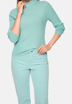 Kvinde iført en lys grønblå ribstrikket sweater og matchende bukser, med den ene hånd rørende ved kraven og den anden hvilende på hoften.