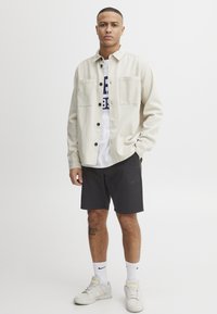 Beige button-up jakke med to frontlommer, hvid t-shirt, sorte shorts, hvide sneakers og hvide sportsokker.