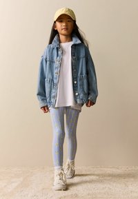 Džínová bunda se zapínáním na knoflíky, bílé tričko, světle modré legíny s květinovým vzorem a béžová čepice. Bílá teniska doplňuje outfit.