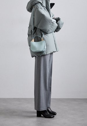 Person iført grå bukser med vide ben, sorte blokhælede støvler, oversized lysegrå dunjakke og bærende en lille blå håndtaske.
