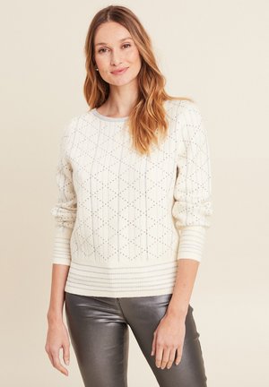 Cremfarbener Pullover mit gerippten Ärmeln und Saum, der mit Rautenmuster und kleinen metallischen Akzenten verziert ist. Getragen mit glanzvoll metallisch grauen Hosen.
