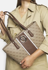 Brązowa torba tote z logo, z wzorzystą jasnobrązową częścią, dwiema długimi rączkami i srebrnymi akcentami.