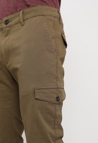 Záběr zblízka na osobu oblečenou v hnědých cargo kalhotách s zapínací kapsou na stehně a vínovým tričkem zastrčeným uvnitř.