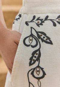 Kreemjas kangas, millel on keerukas must ja pruun lilleline tikand, sisaldades lehe- ja terakujulisi kujundeid. Tasku detail on nähtav.