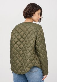 Giacca trapuntata verde oliva con scollatura rotonda, caratterizzata da un motivo a diamante e un orlo leggermente curvo. Materiale morbido e leggero.