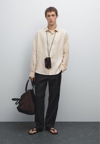 Camicia a maniche lunghe di un beige chiaro, con vestibilità oversize, abbinata a pantaloni neri a gamba larga e infradito neri; borsa in suede marrone scuro e accessori appesi.