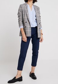 Grau kariertes, doppelt geknöpftes Blazer mit rosa Akzenten, hellblaues Hemd, navy ankle pants und schwarze Slipper. Taillierte Passform, hochgedrehte Ärmel.