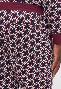 Close-up van een gepatterneerde broek en hand, met een geometrisch ontwerp in marineblauw, bordeauxrood en wit, inclusief een achterzak en een bijpassende tailleband.