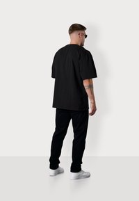 Black oversized t-shirt made of cotton, paired with black jeans. Model stands wearing white sneakers and sunglasses, showing a dragonfly tattoo.